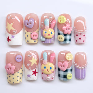 A set of ten artificial nails with a variety of designs. Each nail features a pink base with decorative elements like stars, hearts, buttons, and a cartoon bunny. Some nails have polka dots, stripes, or a checkered pattern.
