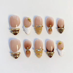 A set of ten artificial nails in various designs. The nails feature a nude base with gold accents, including rings, studs, and decorative elements. Some nails have a gradient effect, while others have a French tip design.