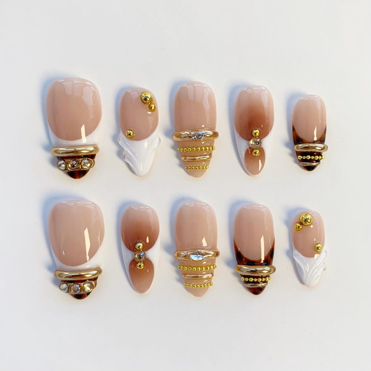 A set of ten artificial nails in various designs. The nails feature a nude base with brown ombre and gold accents. Some nails have gold rings with rhinestones, while others have gold studs and white decorative elements.