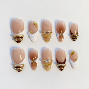A set of ten artificial nails in various designs. The nails feature a nude base with brown ombre and gold accents. Some nails have gold rings with rhinestones, while others have gold studs and white decorative elements.
