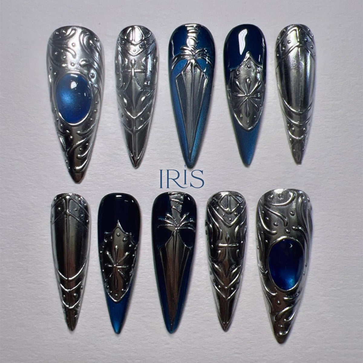 A set of ten long, pointed, silver-colored nail enhancements. Some nails feature a blue gem or a blue and silver design. The word "IRIS" is printed in blue on the surface.