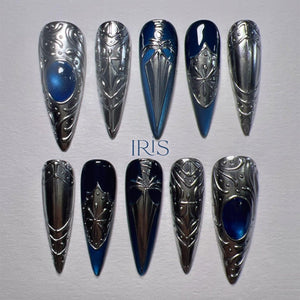 A set of ten long, pointed, silver-colored nail enhancements. Some nails feature a blue gem or a blue and silver design. The word "IRIS" is printed in blue on the surface.