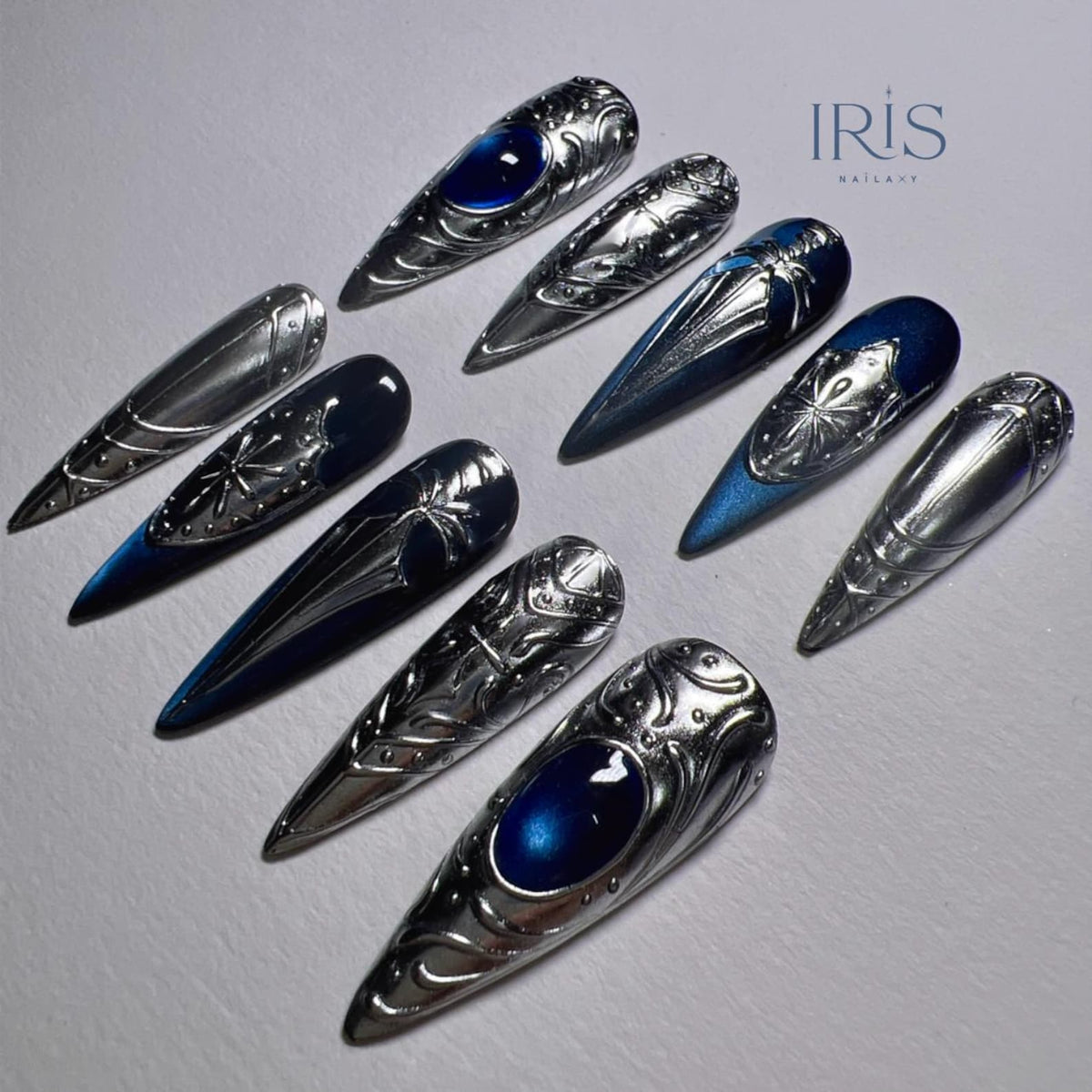 A collection of silver-toned, long, pointed artificial nails. Some nails feature a dark blue gradient and ornate, embossed designs, including a central gem. The brand name "IRIS NAILART" is visible in the upper right corner.