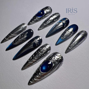 A collection of silver-toned, long, pointed artificial nails. Some nails feature a dark blue gradient and ornate, embossed designs, including a central gem. The brand name "IRIS NAILART" is visible in the upper right corner.