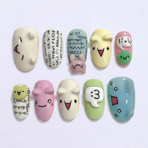 A collection of ten artificial nails, each with a unique design. The nails feature various colors, including white, pink, yellow, green, and blue. Some nails have cartoon faces, symbols, and text. The designs include a bunny, a speech bubble with the