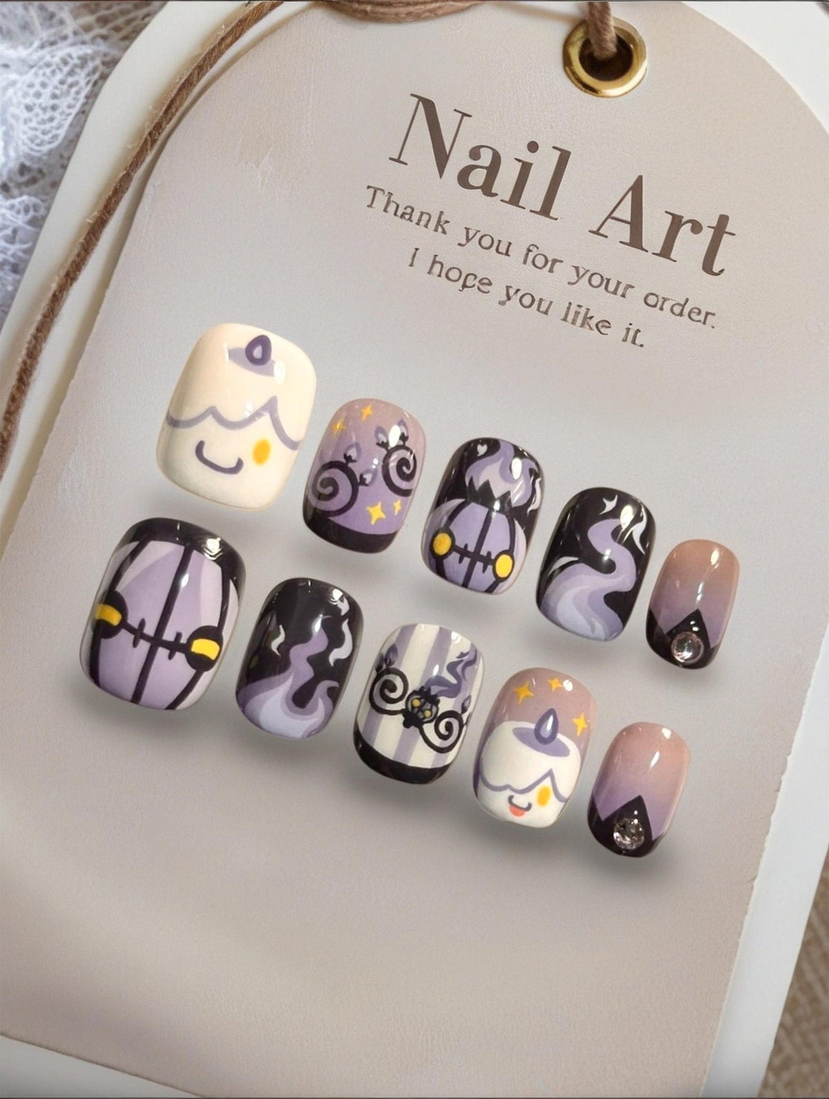 A set of ten press-on nails featuring a Halloween theme with purple, black, and cream colors. Designs include smiling candles, ghosts, pumpkins, and swirling smoke patterns. The display card reads Nail Art Thank you for your order. I hope you like it