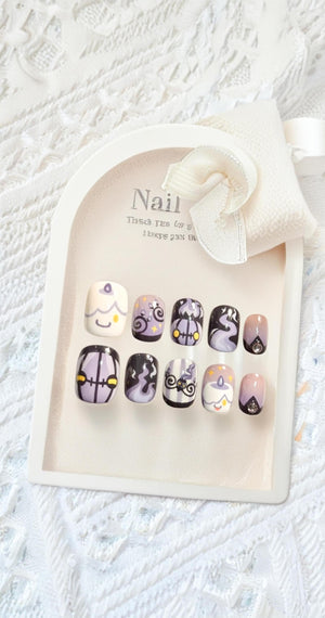 A set of ten press-on nails displayed on a cream-colored arched card with a white ribbon. The nails feature designs including cartoon candles, swirling smoke, and geometric patterns in shades of purple, black, white, and yellow.
