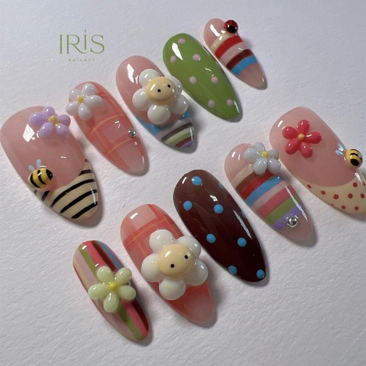 A collection of artificial nails with various designs. Some nails feature 3D flower and bee embellishments. Other nails have striped, polka dot, and plaid patterns. The nails are painted in shades of pink, green, brown, and white.