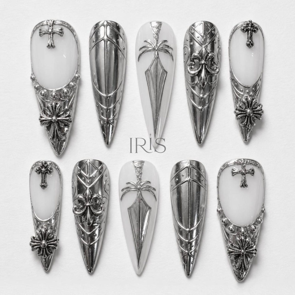 A set of ten silver-toned, long, pointed artificial nails. Each nail features intricate designs, including crosses, fleur-de-lis, and geometric patterns. The nails have a white base and are arranged on a white surface.