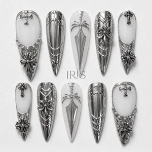 A set of ten silver-toned, long, pointed artificial nails. Each nail features intricate designs, including crosses, fleur-de-lis, and geometric patterns. The nails have a white base and are arranged on a white surface.