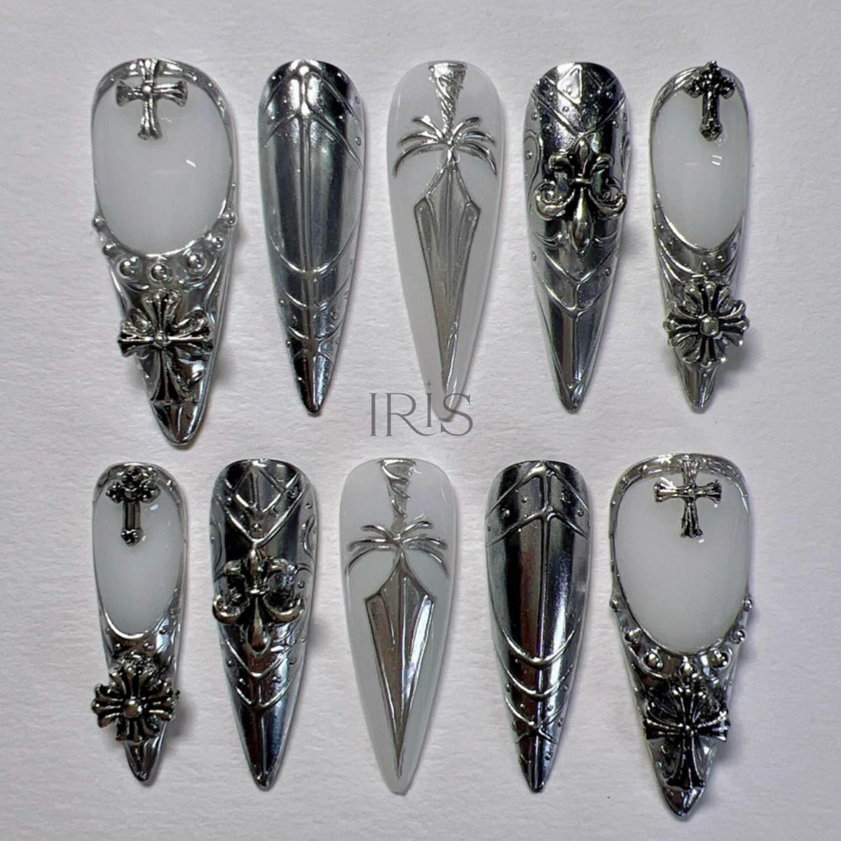 A set of ten long, pointed artificial nails. The nails feature a combination of white and silver designs, including crosses, floral accents, and geometric patterns. The word "IRIS" is visible on one of the nails.
