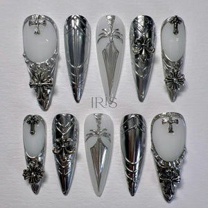 A set of ten long, pointed artificial nails. The nails feature a combination of white and silver designs, including crosses, floral accents, and geometric patterns. The word "IRIS" is visible on one of the nails.