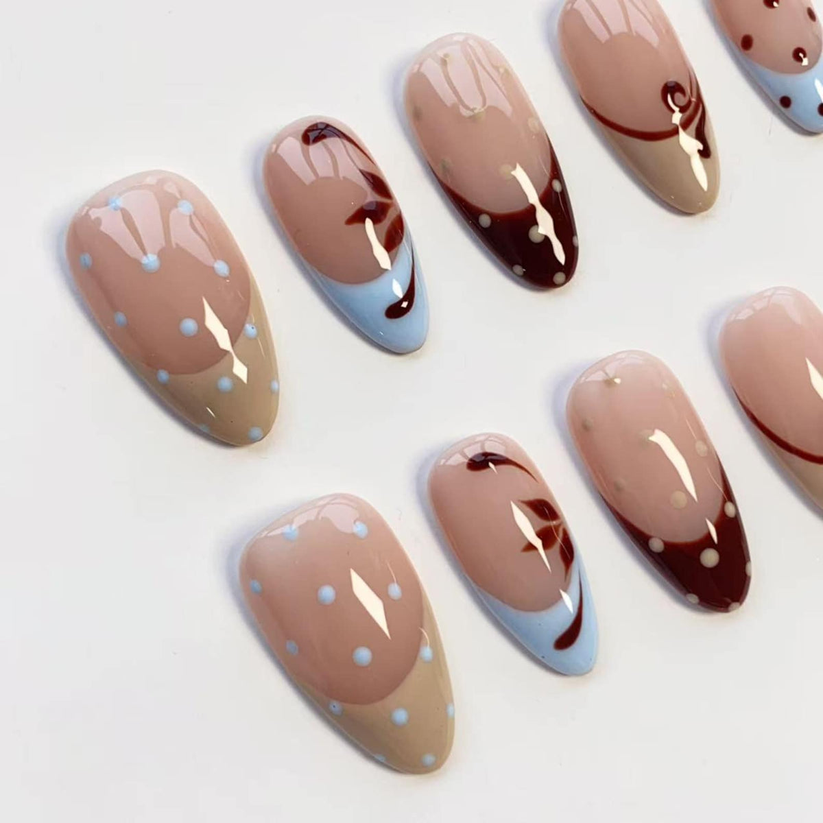 A collection of oval-shaped artificial nails. Some nails feature a light brown base with a light blue tip and a dark brown floral design. Others have a light brown base with light blue polka dots.