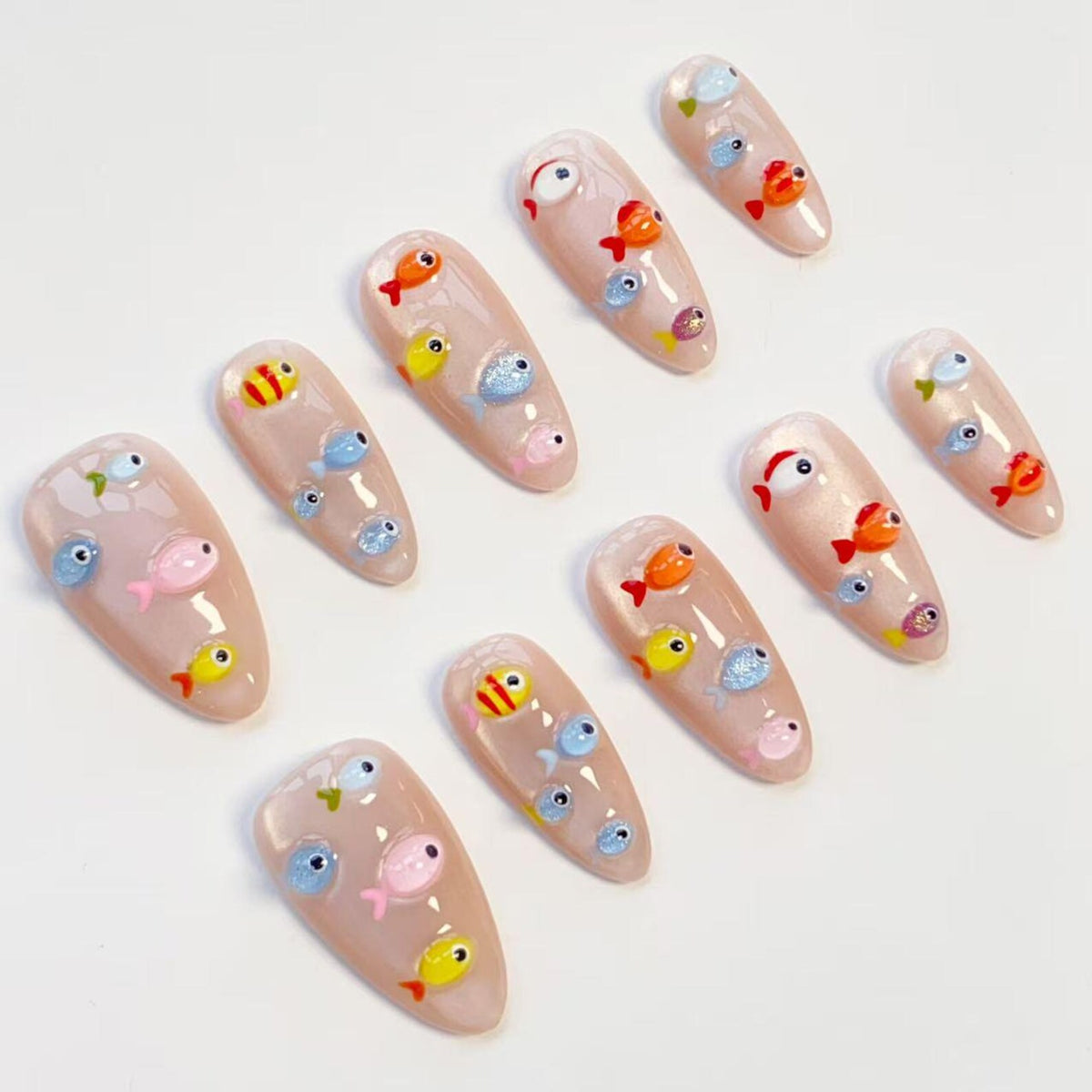 A set of almond-shaped artificial nails, each adorned with colorful, miniature fish designs. The fish are in shades of orange, yellow, blue, and pink, set against a nude-colored base. The nails are arranged on a white surface.