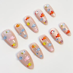 A set of almond-shaped artificial nails, each adorned with colorful, miniature fish designs. The fish are in shades of orange, yellow, blue, and pink, set against a nude-colored base. The nails are arranged on a white surface.