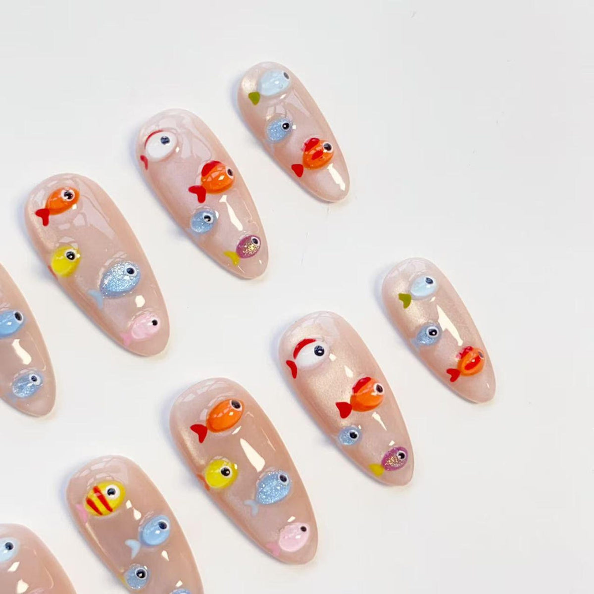A collection of almond-shaped artificial nails, each painted with a nude base and adorned with colorful, cartoonish fish designs. The fish are in various colors, including orange, yellow, blue, and pink, with detailed eyes and fins.