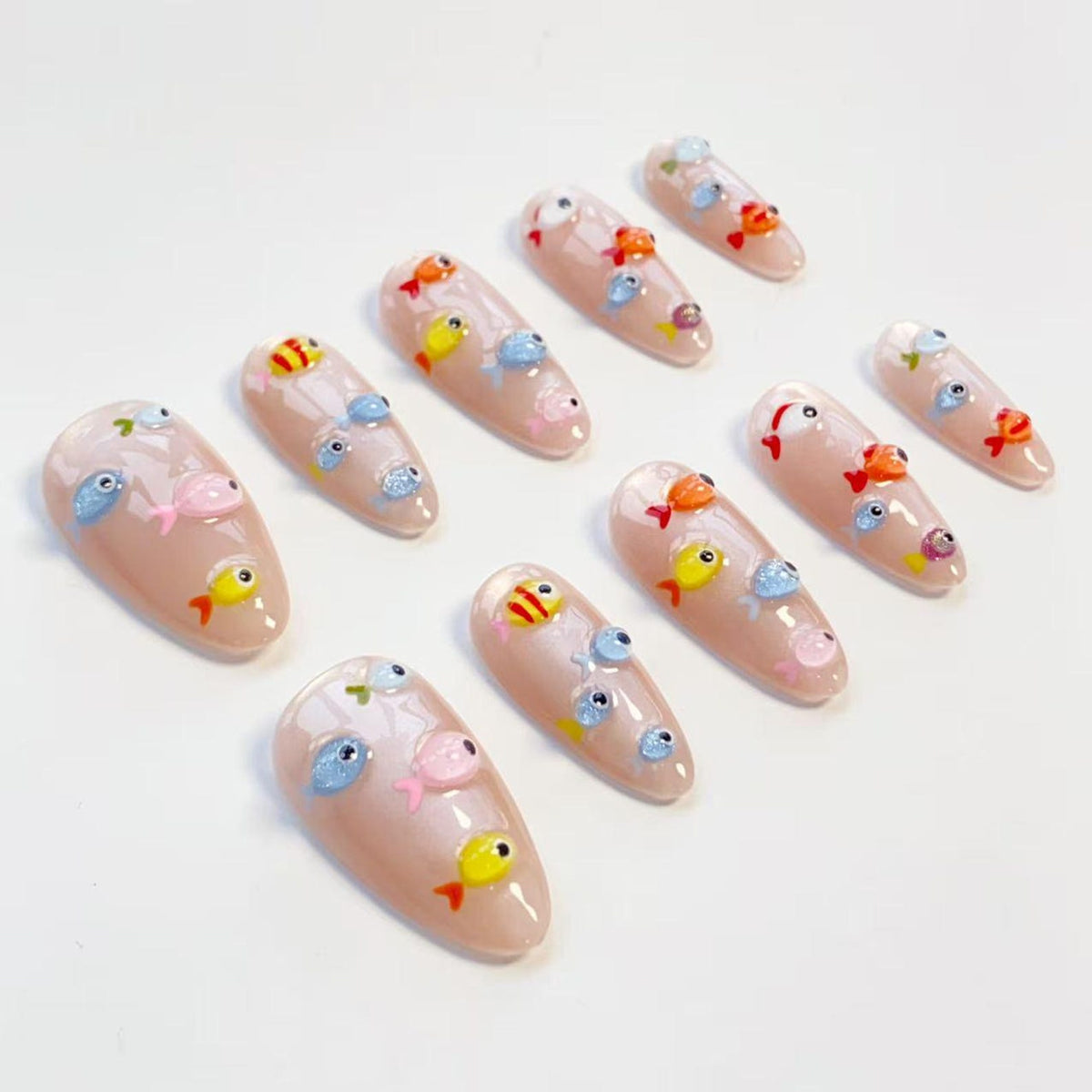 A set of almond-shaped false nails with a nude base color. Each nail is decorated with colorful, miniature fish designs in shades of yellow, orange, pink, and blue. The fish are arranged in various positions, creating a playful, aquatic theme.