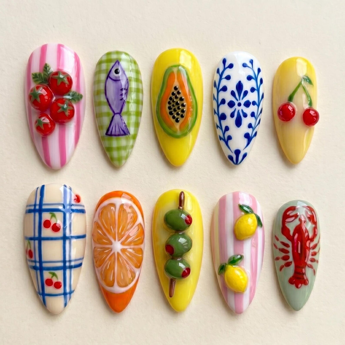A set of ten artificial nails with various food-themed designs. The designs include tomatoes, a fish, papaya, cherries, a blue floral pattern, cherries, an orange slice, olives, lemons, and a lobster. Each nail has a different color and design.