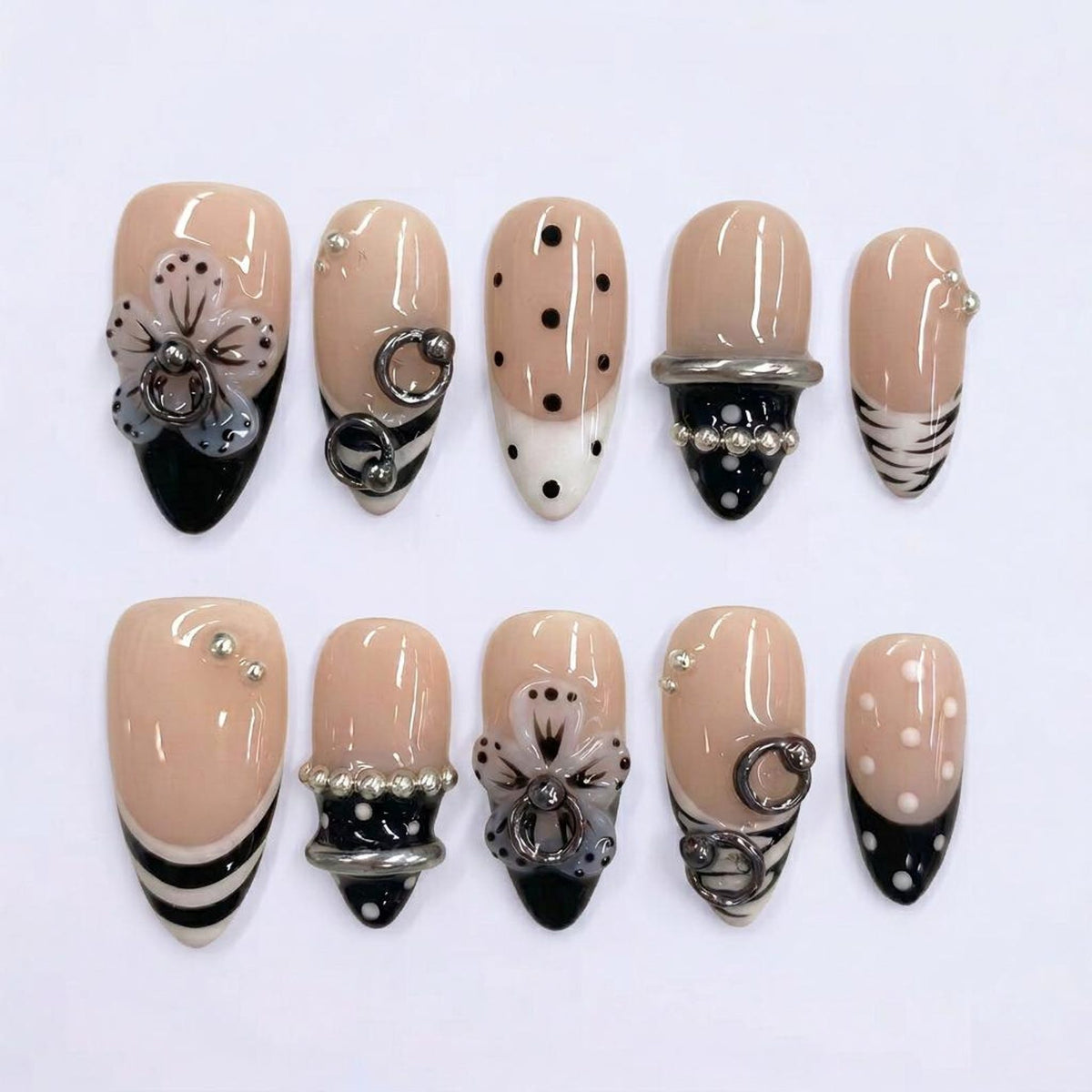 A set of ten almond-shaped artificial nails. The nails feature a nude base with various designs, including black tips, polka dots, floral accents, and silver embellishments. Some nails have silver rings and beaded details.