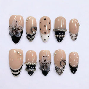 A set of ten almond-shaped artificial nails. The nails feature a nude base with various designs, including black tips, polka dots, floral accents, and silver embellishments. Some nails have silver rings and beaded details.