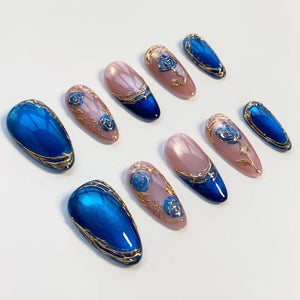 A set of ten artificial nails in various designs. Some nails are solid blue with gold trim, while others feature a pink base, blue tips, and gold floral accents. The nails are arranged on a white surface.