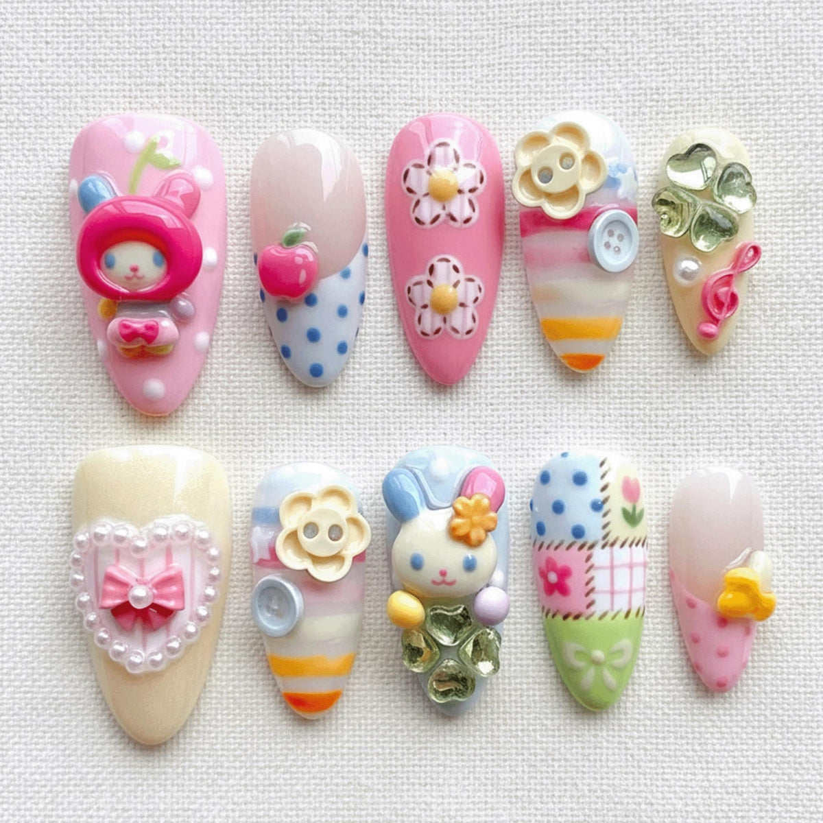 A collection of ten artificial nails, each uniquely decorated with 3D embellishments. Designs include a bunny, flowers, an apple, a heart, and polka dots, in a palette of pink, blue, yellow, and white. The nails are oval-shaped.