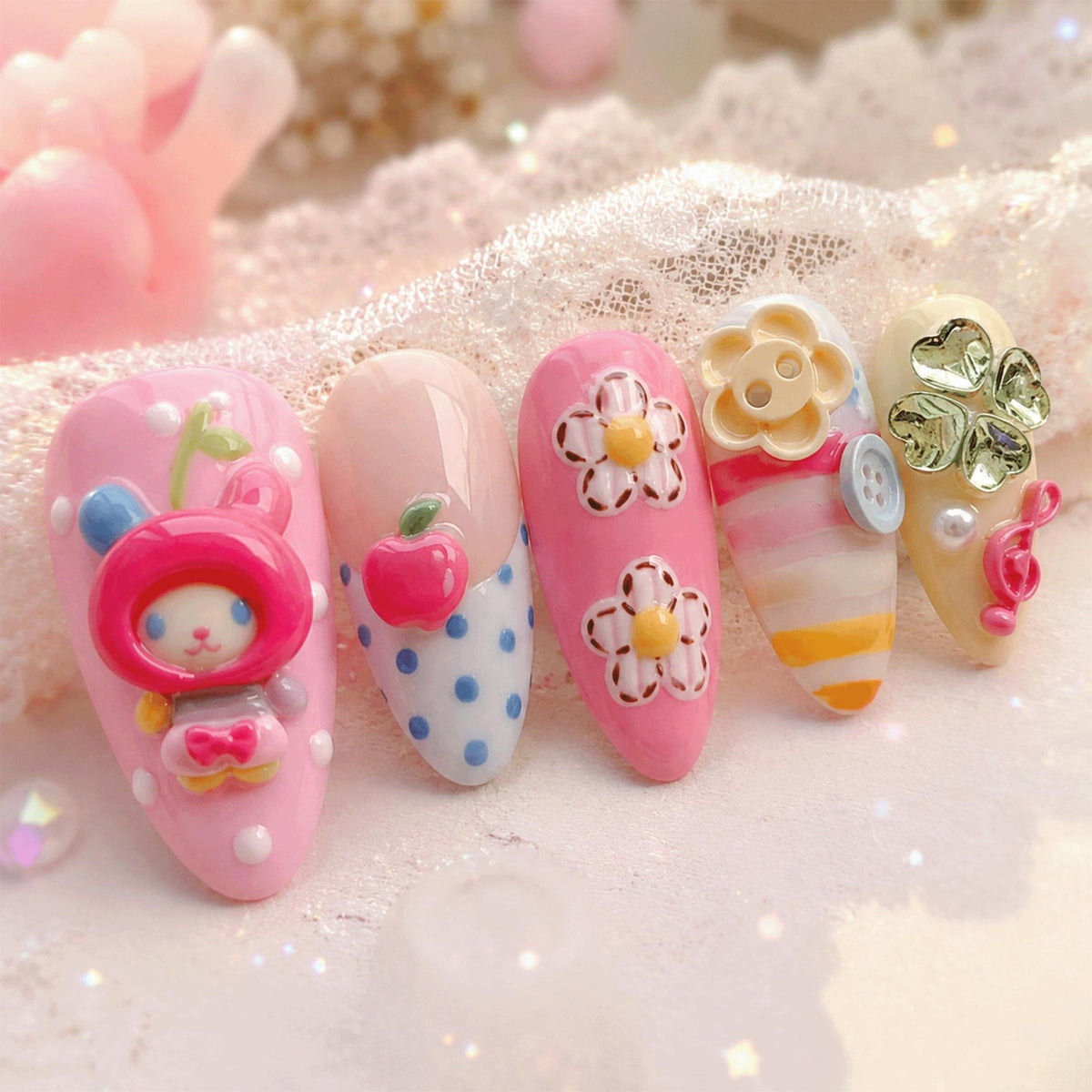A set of five artificial nails with various designs. One nail features a pink bunny, another has a red apple, and two others display white flowers on a pink background. The last nail has a flower, button, and striped design.