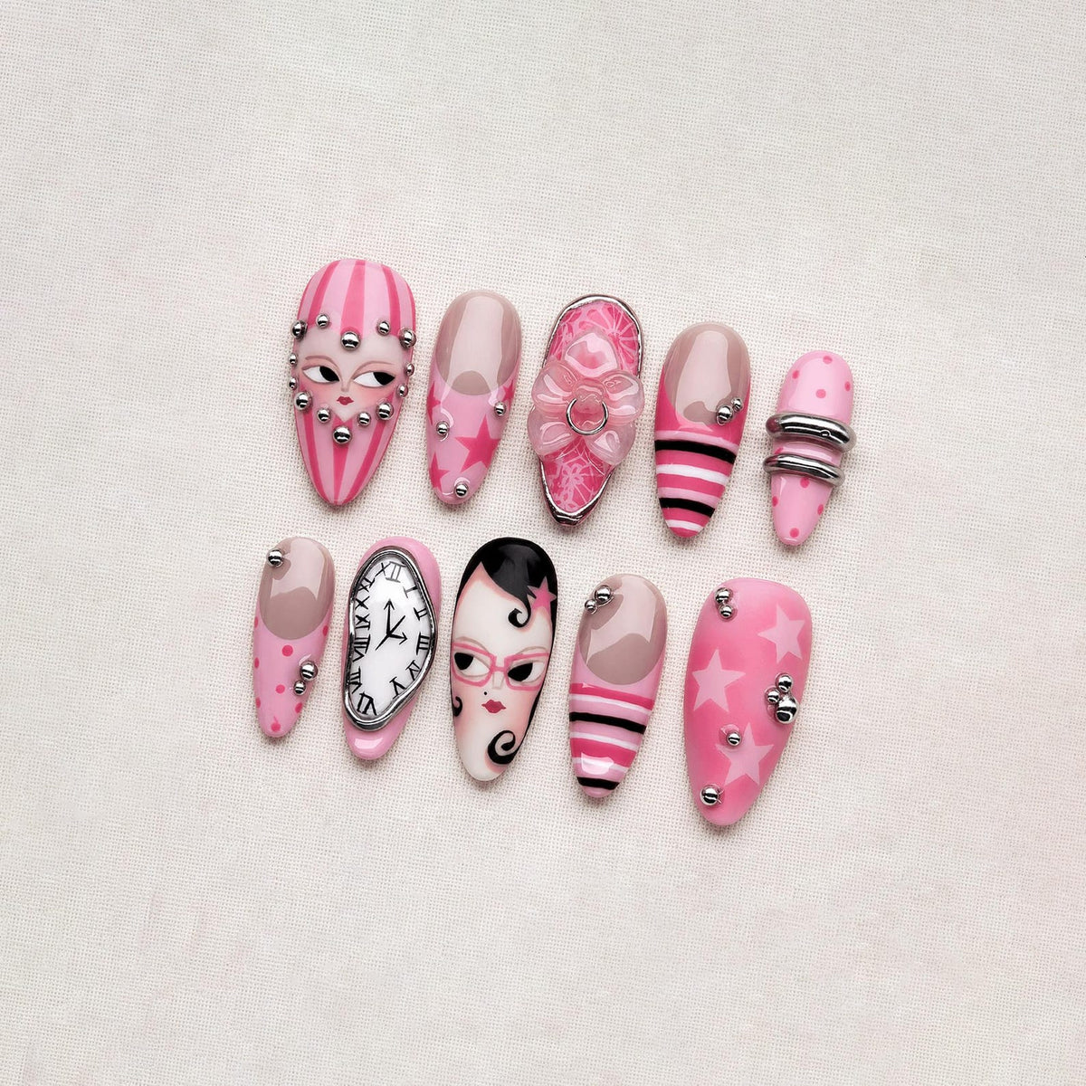 A collection of ten pink and nude-colored artificial nails with various designs. Some nails feature faces, stars, stripes, and silver embellishments. One nail displays a clock face, while another has a floral design.