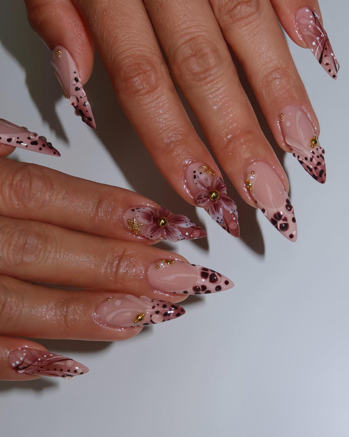 Long, pointed acrylic nails with a nude base, adorned with dark brown polka dots, floral designs, and gold accents. Some nails feature a gradient effect and delicate white lines, creating a sophisticated and artistic manicure.