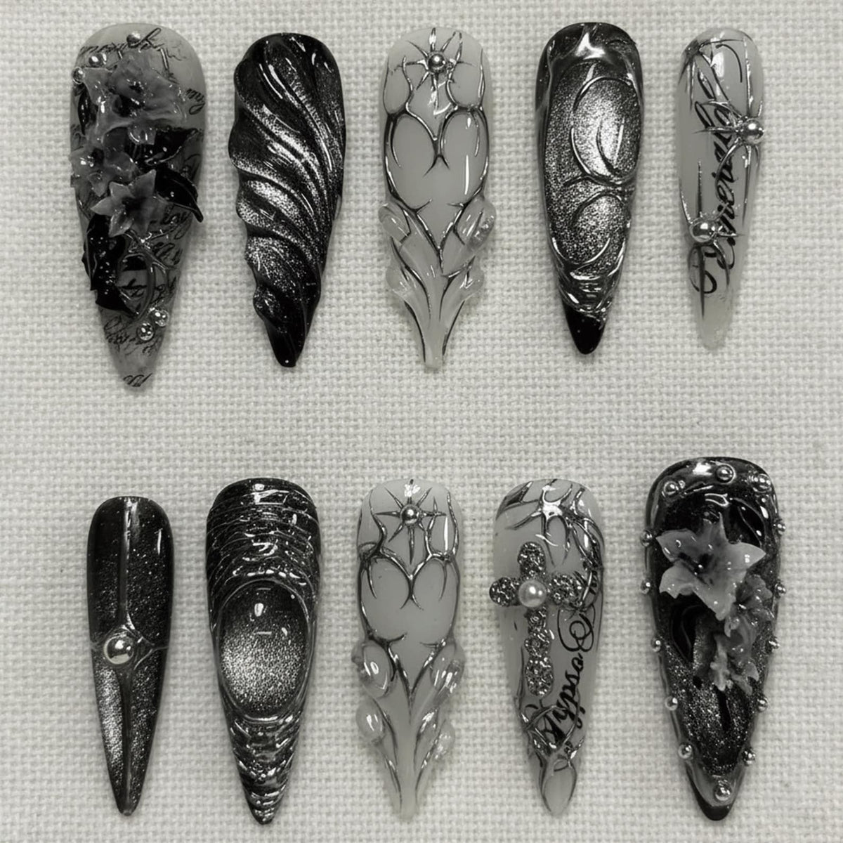 A set of ten long, pointed acrylic nails with intricate designs. The nails feature a variety of embellishments, including silver accents, floral patterns, and text. The color palette is primarily black, white, and silver, creating a gothic aesthetic.