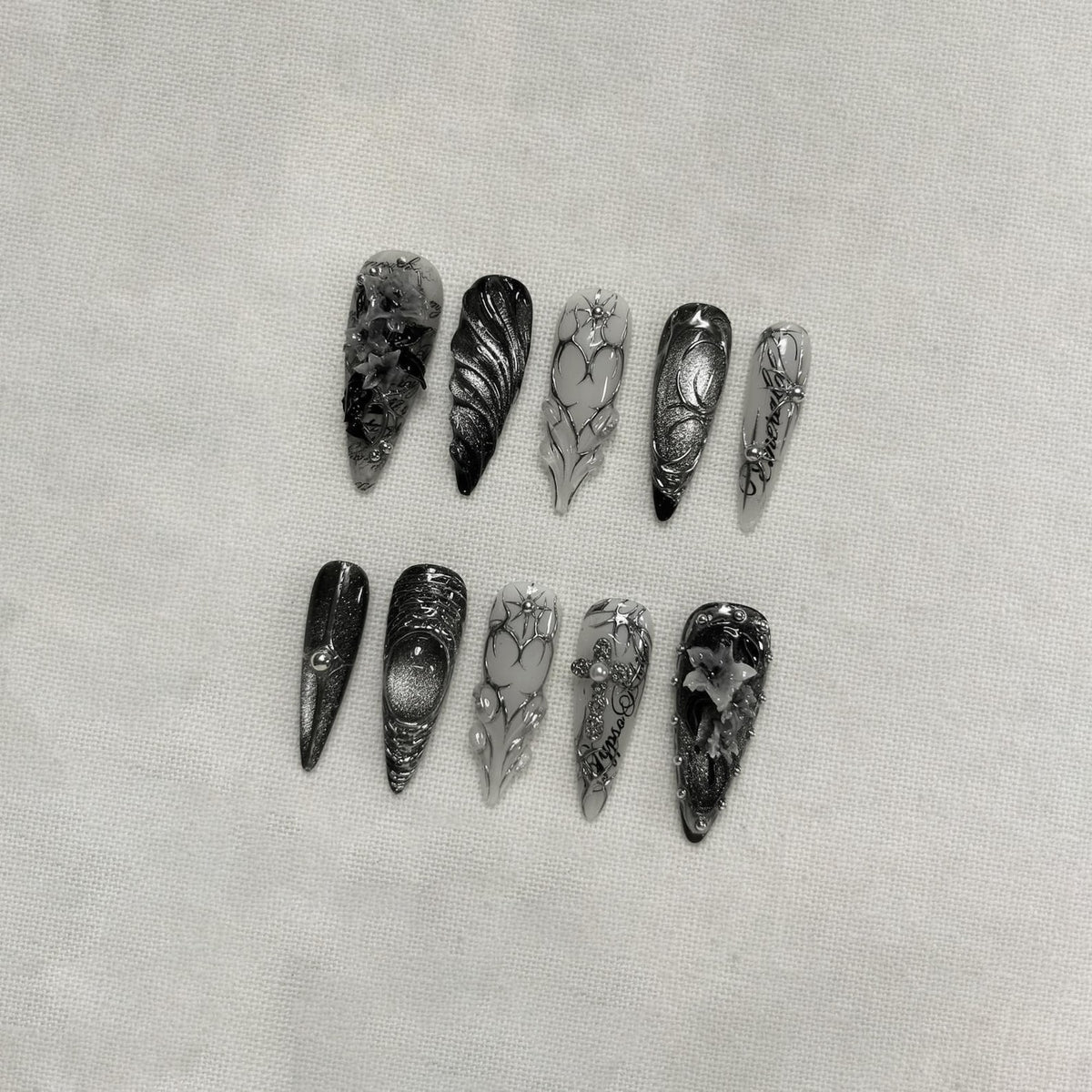 A collection of ten long, almond-shaped artificial nails. Each nail features a unique design in shades of black, gray, and silver. Some nails have floral patterns, while others have abstract designs or embellishments.