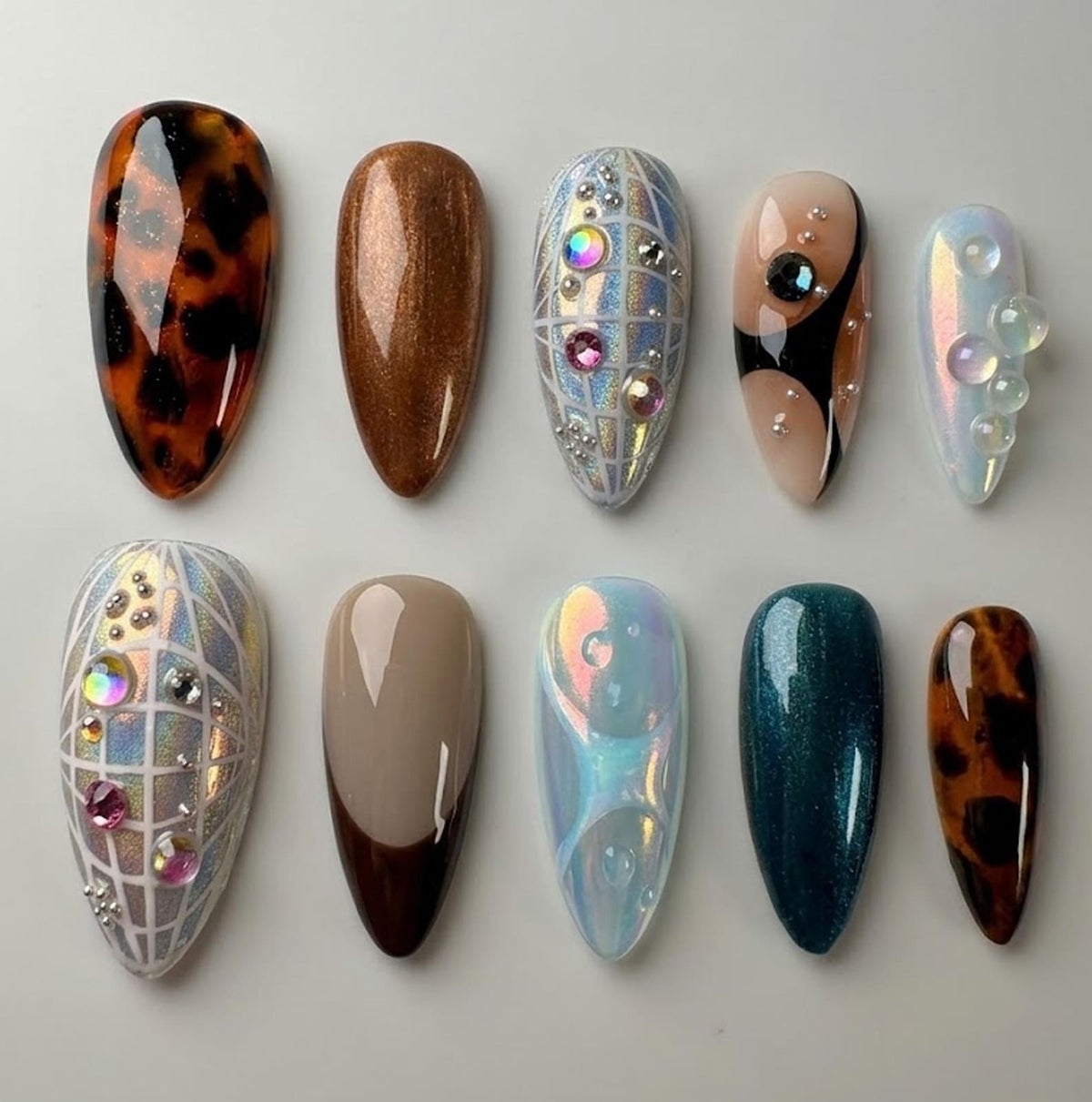 An assortment of ten almond-shaped artificial nails. The nails feature various designs, including tortoiseshell, glitter, iridescent, and geometric patterns. Some nails are adorned with rhinestones and other embellishments. The color palette includes