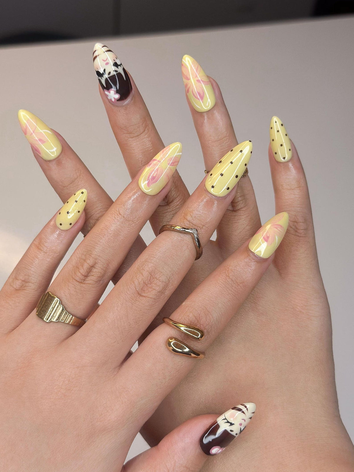 A set of long, almond-shaped nails with various designs. Some nails are yellow with pink floral patterns or black polka dots. Others feature a brown and white design with a floral accent. Gold rings adorn the fingers.