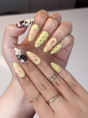 A hand displays long, almond-shaped nails with various designs. Some nails are yellow with pink floral patterns or black polka dots. Other nails feature a black and white design with floral accents. Gold rings adorn the fingers.