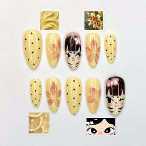A set of ten artificial nails in various designs. Some nails are yellow with black polka dots, others feature floral patterns in pink and orange, and two nails have cat face designs. Additional images include a close-up of a flower, lemon slices, and