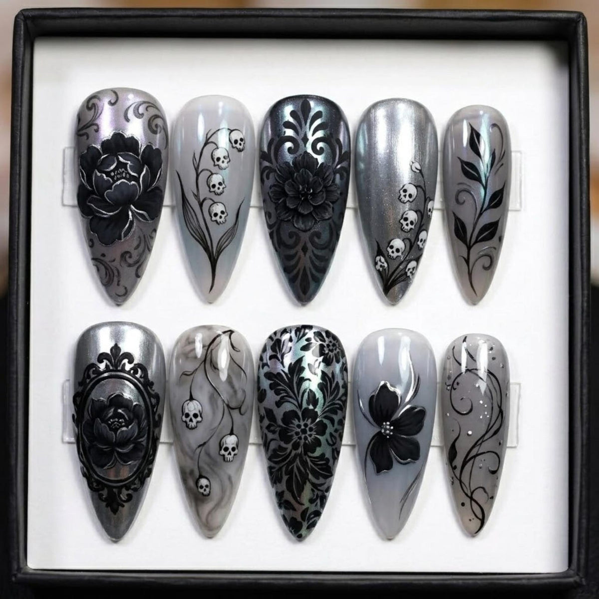 A set of ten long, pointed, artificial nails with intricate designs. The nails feature a variety of black floral and skull patterns on a silver and gray background. The designs include roses, damask, and skull accents.