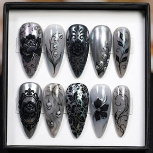A set of ten long, pointed, artificial nails with intricate designs. The nails feature a variety of black floral and skull patterns on a silver and gray background. The designs include roses, damask, and skull accents.