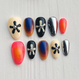 A set of ten artificial nails in various designs. The nails feature a mix of colors, including cream, black, silver, and orange. Some nails have a black flower design with a silver gem, while others have a gradient effect.