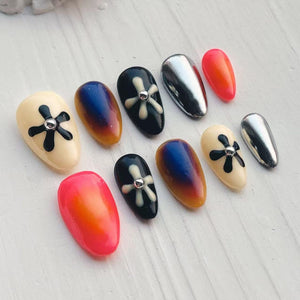 A collection of ten artificial nails in various designs. The nails feature a range of colors, including cream, black, silver, and vibrant orange. Some nails have a flower design, while others have a gradient effect.