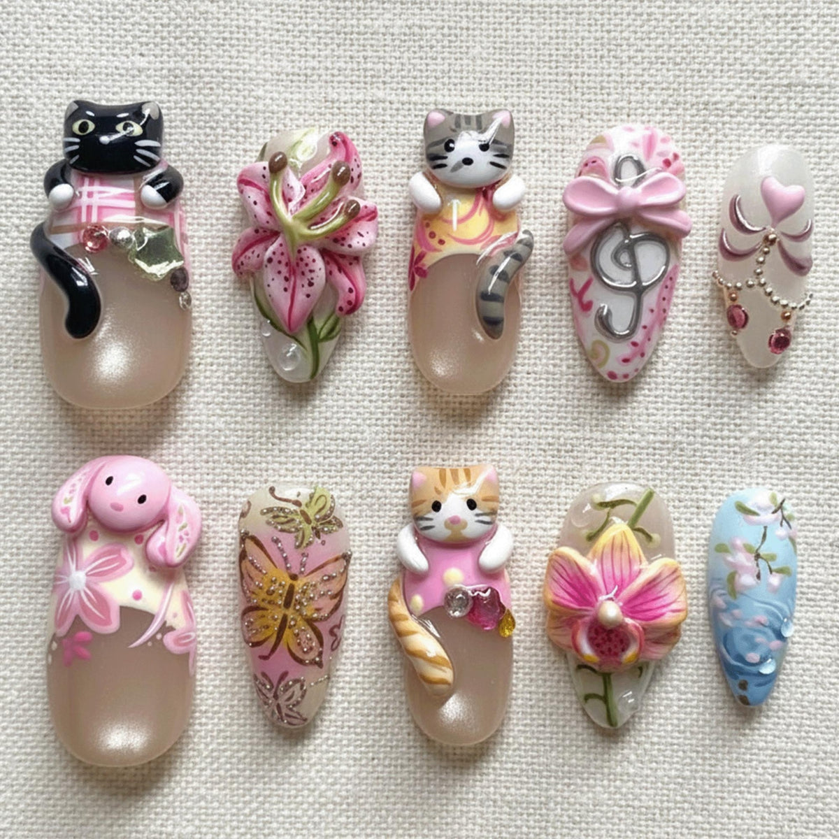 A collection of ten nail art designs. The designs feature 3D elements like cats, flowers, and butterflies. Colors include pink, white, and beige. Some nails have bows and decorative accents.