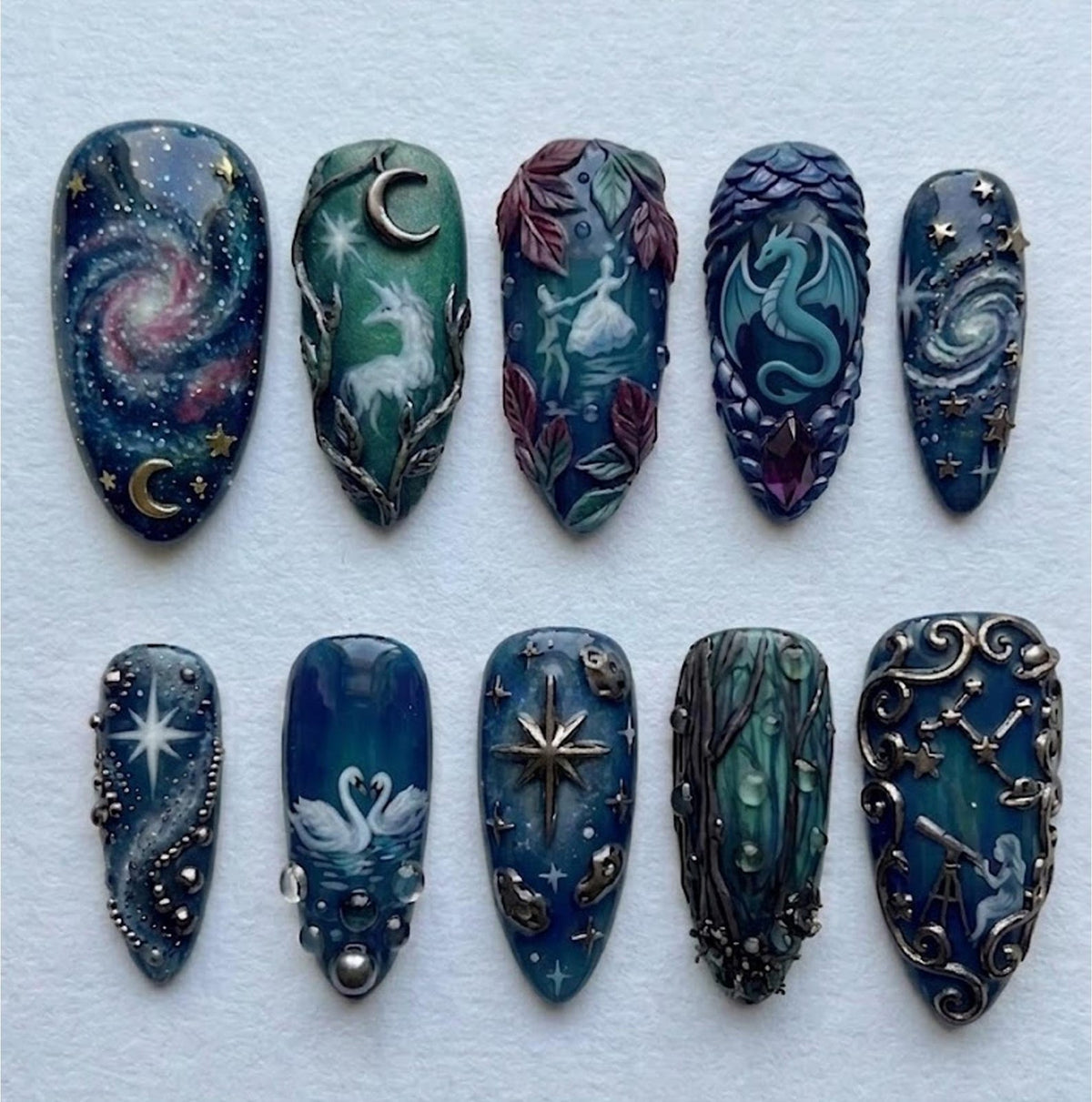 A set of ten intricately designed artificial nails. Each nail features a unique, hand-painted design with celestial and fantasy themes, including galaxies, dragons, and constellations. The color palette includes blues, greens, and metallic accents.