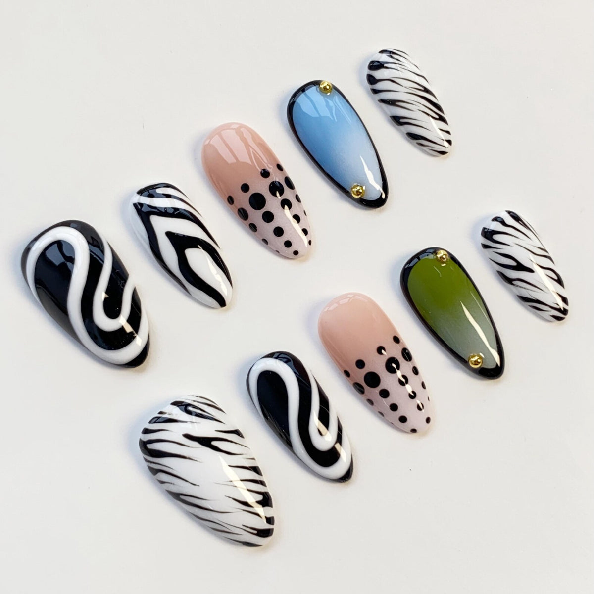 A collection of ten artificial nails in various designs. The nails feature patterns such as black and white zebra stripes, black swirls on white, and nude with black polka dots. Some nails have a light blue or olive green base with gold accents.