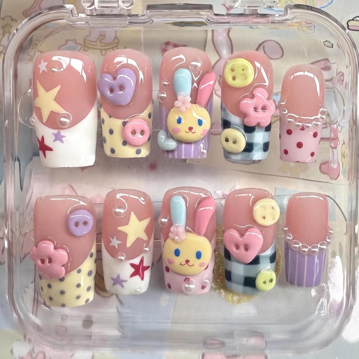 A set of ten press-on nails in a clear plastic case. The nails feature a variety of designs, including stars, hearts, buttons, and a cartoon bunny. The nails are primarily pink, white, yellow, and blue.