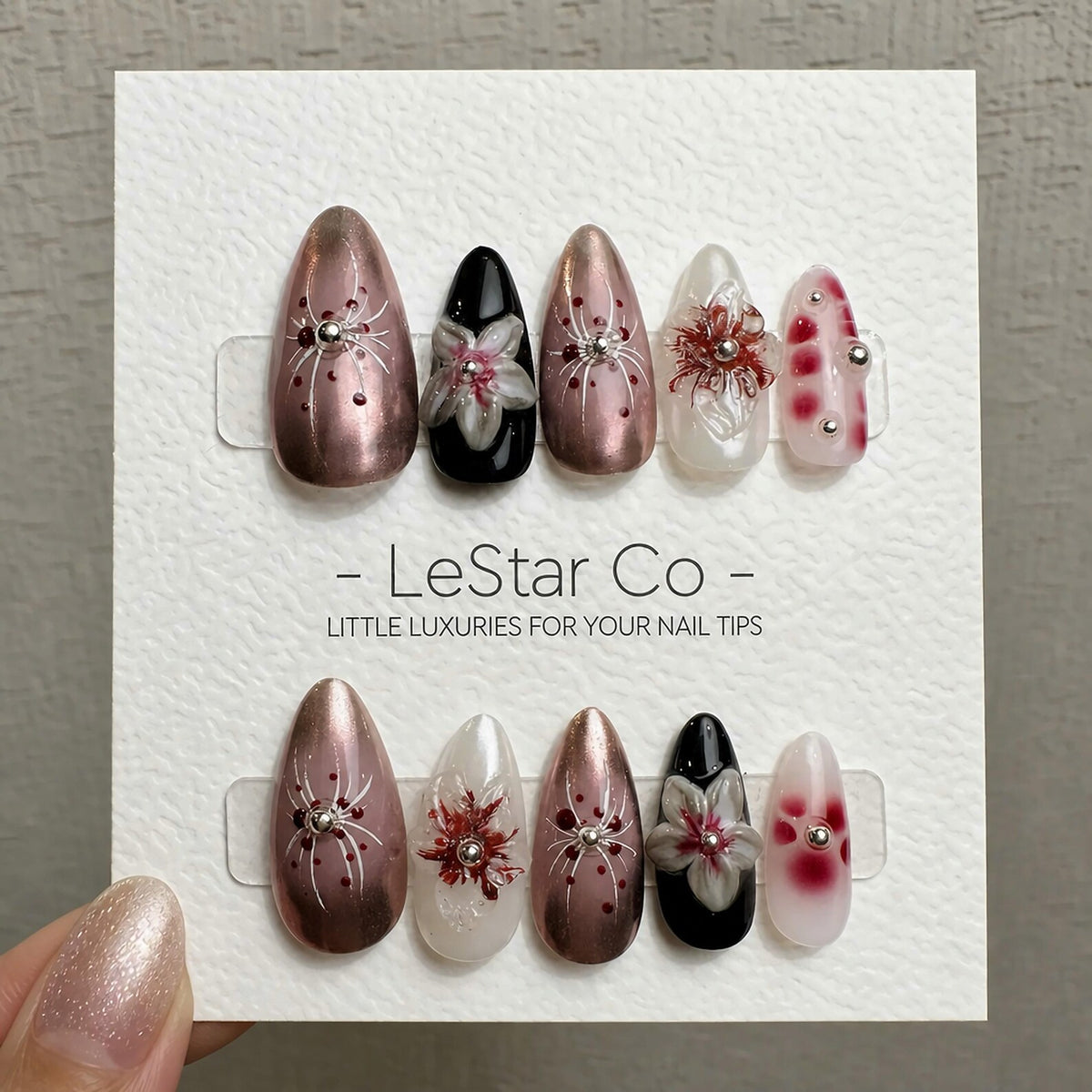 A set of almond-shaped false nails with various designs. The nails feature metallic pink, black, and white bases, adorned with floral and spiderweb-like patterns. Some nails have silver embellishments