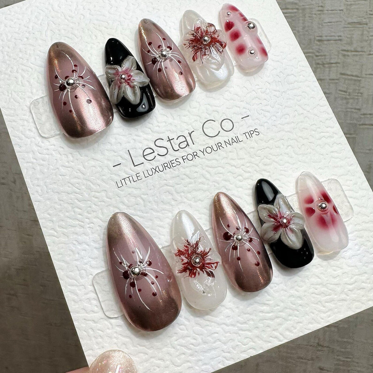 A set of artificial nails in various designs. Some nails are metallic rose gold with a spiderweb design, others are black with a white flower, and some are clear with red floral accents. The text "- L