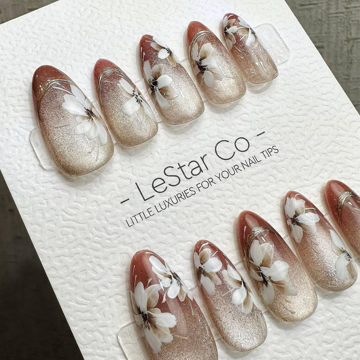 Set of artificial nails with a floral design. The nails have a gradient effect, transitioning from a reddish-brown at the cuticle to a shimmering gold at the tip. Each nail features a white floral pat