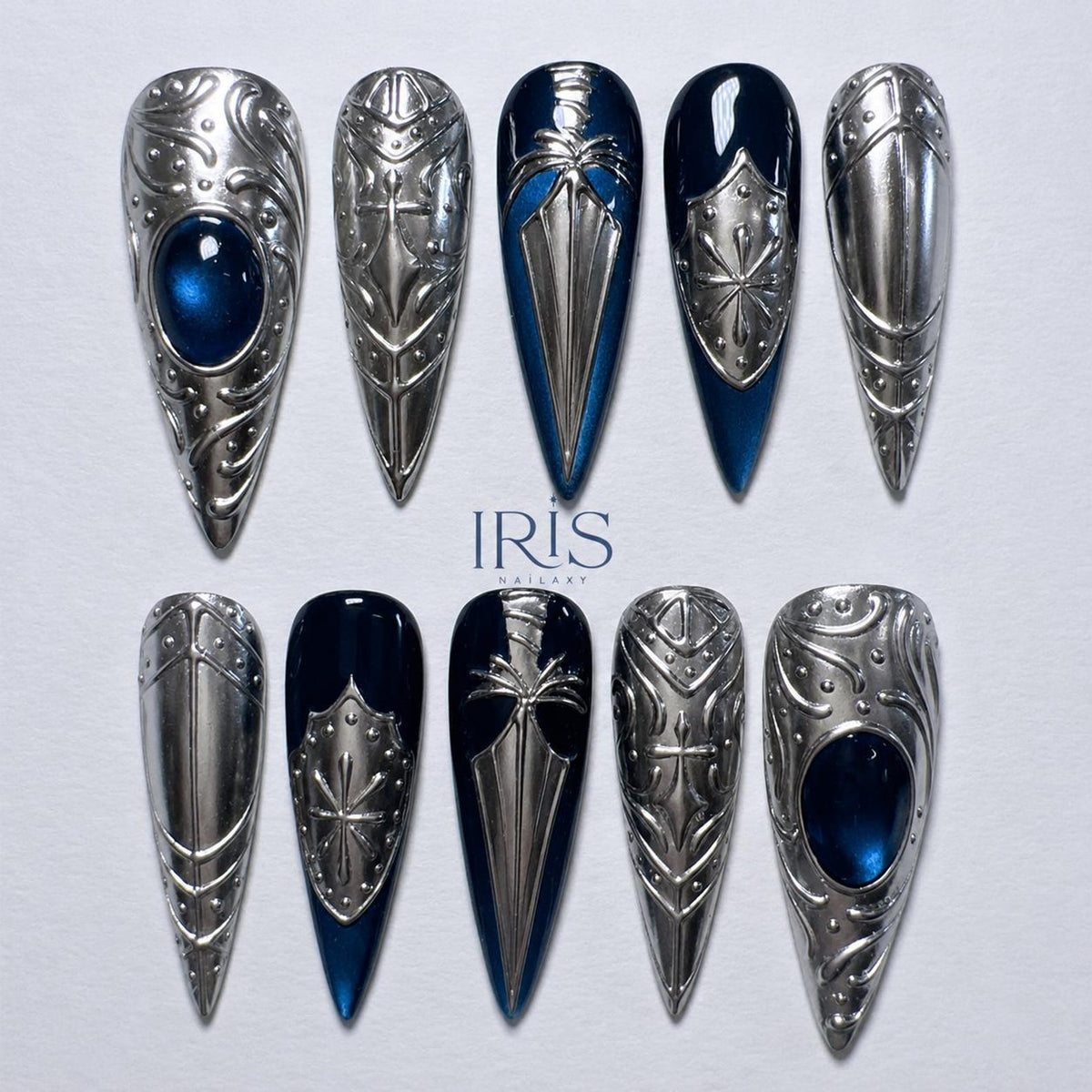 A set of ten long, pointed, silver-colored nail designs. Some nails feature a dark blue base with silver embellishments, while others are entirely silver with a blue gem. The text "IRIS NAILARY" is visible.