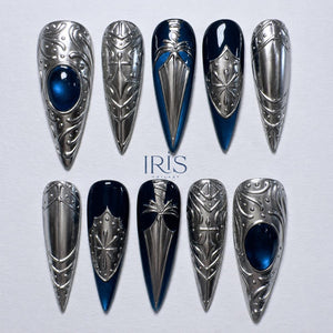A set of ten long, pointed, silver-colored nail designs. Some nails feature a dark blue base with silver embellishments, while others are entirely silver with a blue gem. The text "IRIS NAILARY" is visible.