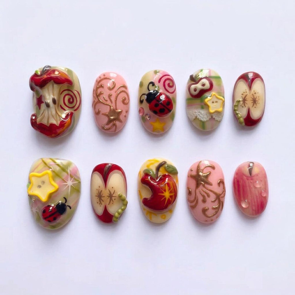 A set of ten artificial nails with various designs. The nails feature detailed artwork including apples, ladybugs, stars, and swirls in shades of red, pink, yellow, and gold. Each nail has a unique, hand-painted design.