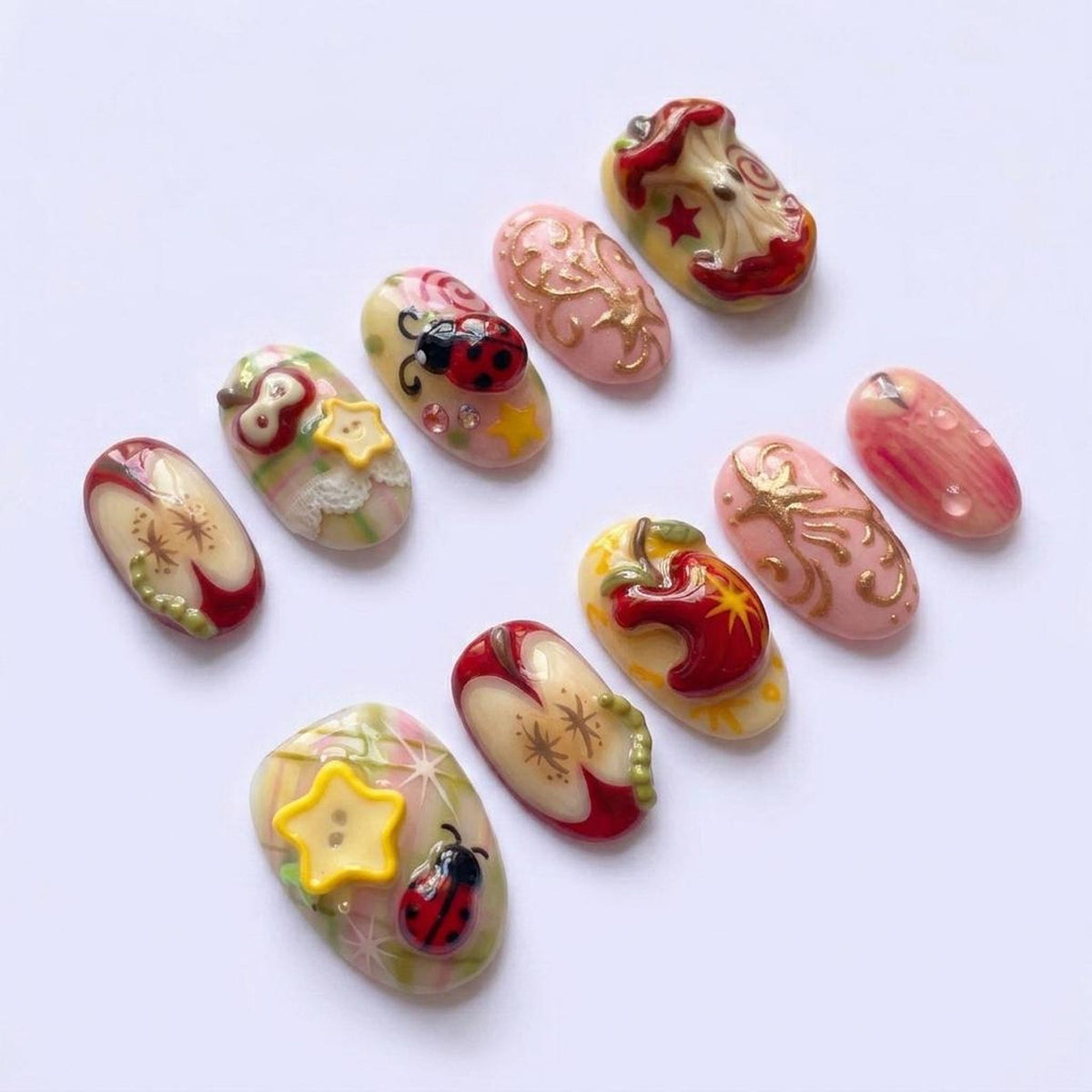 A collection of elaborately decorated artificial nails. Designs include red apples, ladybugs, gold filigree, and yellow stars. The nails are oval-shaped and feature a variety of colors, including red, pink, yellow, and green. The nails are arranged o